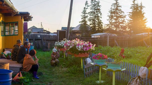 Tres ancianos Yakuts y una joven asiática están charlando en el umbral de una casa de pueblo en el jardín entre macizos de flores en los rayos del sol poniente
. - Foto, imagen