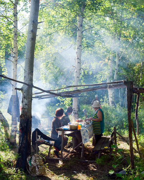 Пожилые якуты накрыли стол в месте отдыха в диком северном лесу в дыму от огня в лучах солнца, падающих сквозь деревья
. - Фото, изображение