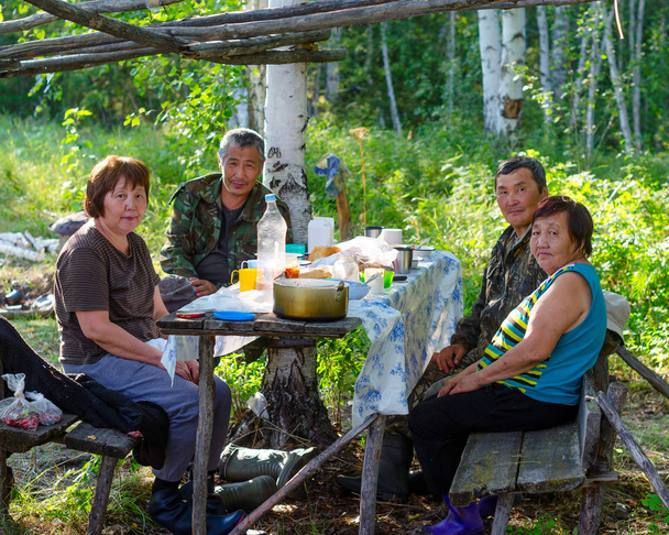 Idősebb ázsiai nők és férfiak portréja családi lakoma az északi Yakut erdőben délután a tűz füstje mögött, múló étellel. - Fotó, kép
