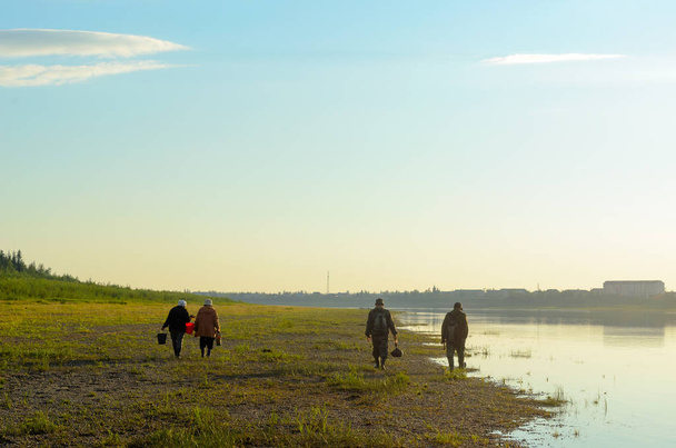 eine Gruppe von Männern und Frauen asiatischer Yakuten mit Säcken und Eimern, die in der Sonne am steinernen wilden Ufer des nördlichen Flusses vilyu und den Häusern des Dorfes ulus suntar bei Sonnenuntergang spazieren gehen. - Foto, Bild