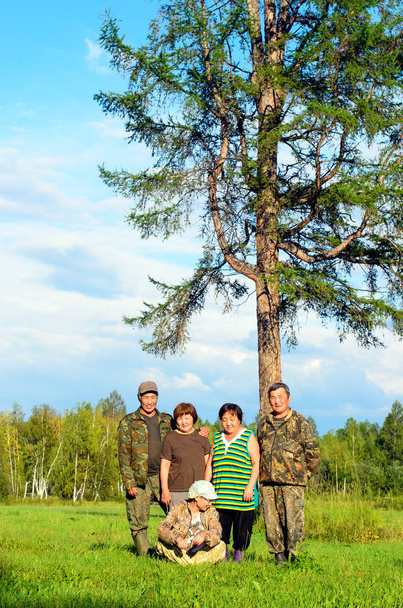 Dos Yakut Asiático ancianos parejas hombres y mujeres con una joven sentada en la hierba posan para una foto de familia en un campo cerca de un gran árbol solitario
. - Foto, Imagen