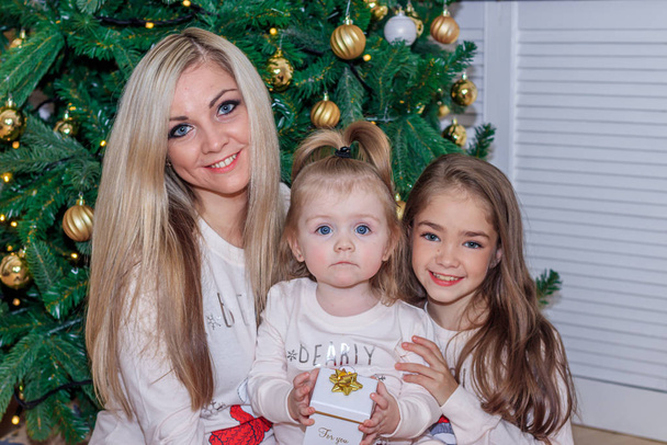 Μαμά και κόρες στο χριστουγεννιάτικο δέντρο. Χριστούγεννα. Αδερφές. Καλά Χριστούγεννα και καλές γιορτές. Χαρούμενη μαμά και οι χαριτωμένες κόρες της ανταλλάσσουν δώρα.. - Φωτογραφία, εικόνα