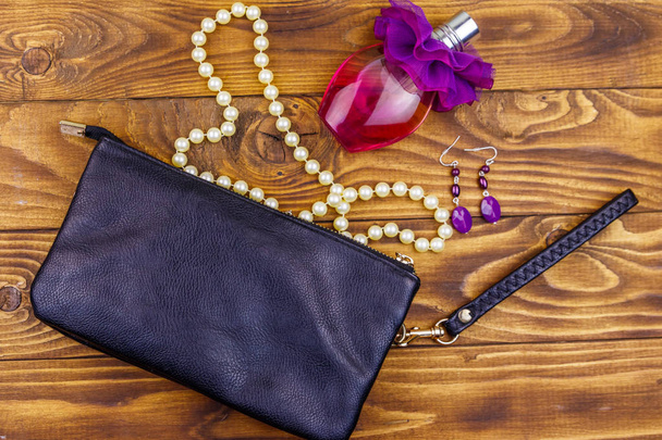 Women accessories on wooden background. Clutch bag, bottle of perfume, pearl necklace and earrings on wood table. Beauty and fashion composition. Top view, flat lay - Photo, Image