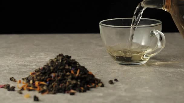 茶道。緑茶は、緑茶の葉のヒープの近くに黒の背景にテーブルの上に透明なマグカップに注がれます - 映像、動画