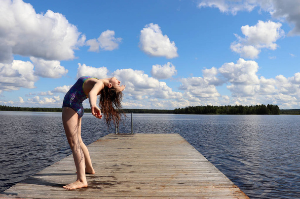 Teenage girl (14) bending over backwards on a wooden dock at a Lake Ranuanjarvi in Ranua, Lapland, Finland on a warm cloudy summer day.   - Fotó, kép