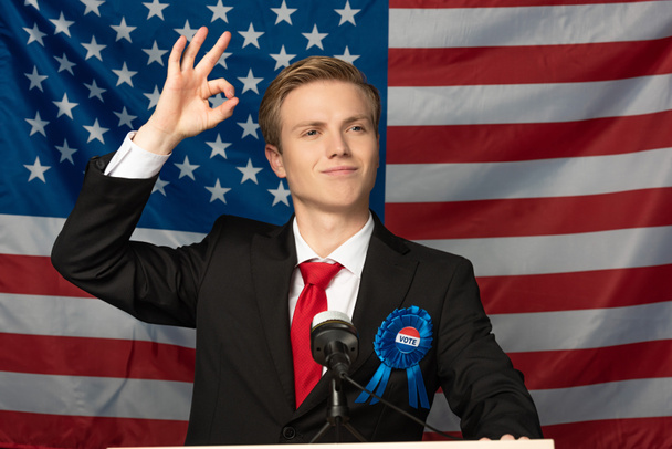 lächelnder Mann zeigt Ok-Zeichen, während er auf der Tribüne auf amerikanischem Flaggenhintergrund steht - Foto, Bild
