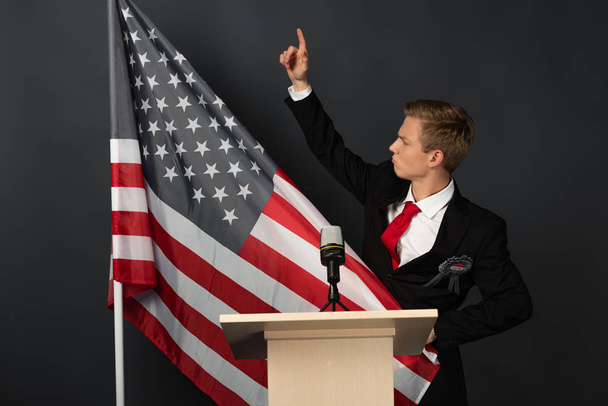 недовольный эмоциональный человек с поднятой рукой на трибуне с американским флагом на черном фоне
 - Фото, изображение