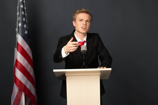 Emotionaler Mann spricht auf Tribüne mit amerikanischer Flagge auf schwarzem Hintergrund - Foto, Bild