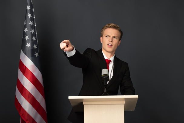 Emotionaler Mann zeigt mit Finger auf Tribüne mit amerikanischer Flagge auf schwarzem Hintergrund - Foto, Bild