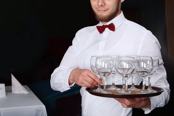 Молодий чоловік-офіціант приніс келихи для вина в лоток. Фото без обличчя в інтер'єрі ресторану
. - Фото, зображення