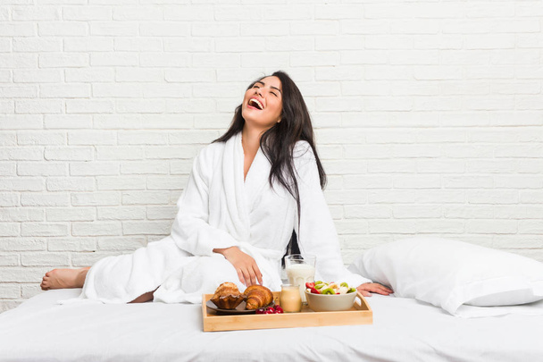 junge kurvige Frau frühstückt entspannt und glücklich lachend auf dem Bett, der Hals gestreckt und zeigt Zähne. - Foto, Bild