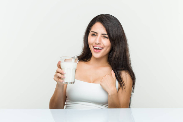 Jeune femme hispanique tenant un verre de lait surprise se montrant du doigt, souriant largement
. - Photo, image