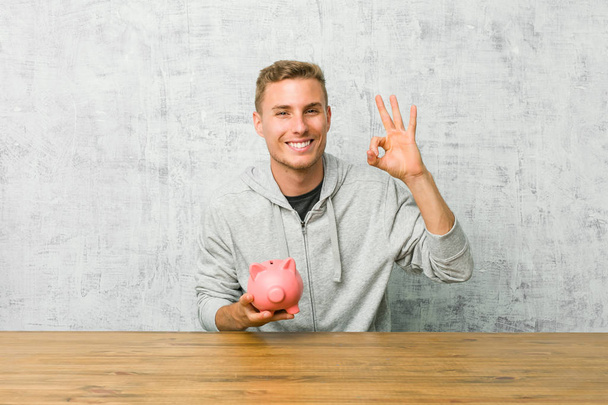 Jovem economizando dinheiro com um banco porquinho alegre e confiante mostrando gesto ok
. - Foto, Imagem