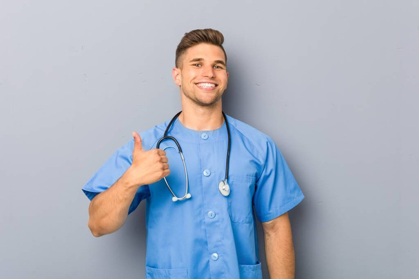Νεαρός νοσοκόμος χαμογελάει και σηκώνει τον αντίχειρα - Φωτογραφία, εικόνα