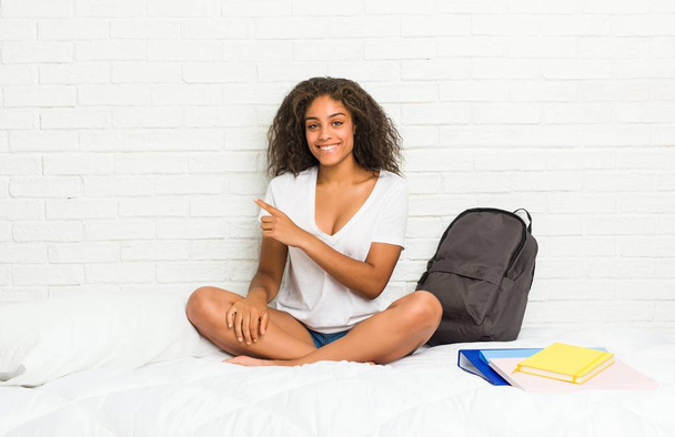 junge afrikanisch-amerikanische Studentin auf dem Bett, lächelnd und zur Seite zeigend, etwas im leeren Raum zeigend. - Foto, Bild