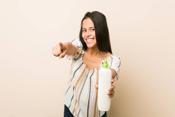 Jeune femme hispanique tenant une bouteille de crème sourire joyeux pointant vers l'avant
. - Photo, image