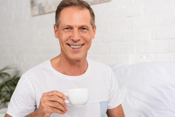 Homem segurando xícara de café e sorrindo para a câmera no quarto
 - Foto, Imagem