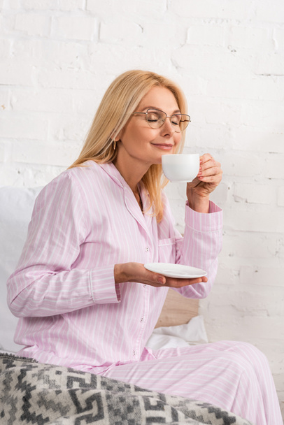 Smiling woman in pajamas smelling coffee on bed - Фото, зображення