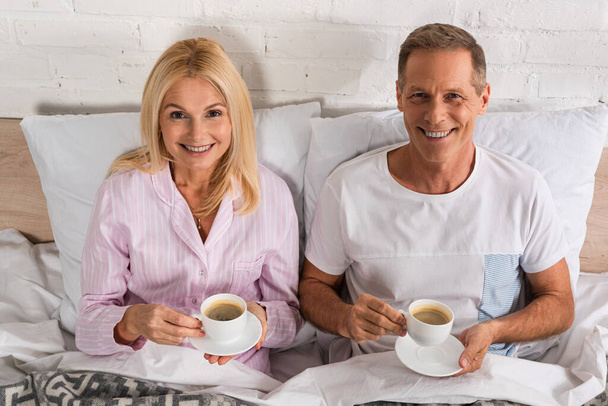 Couple with coffee cups smiling at camera in bed - Фото, изображение