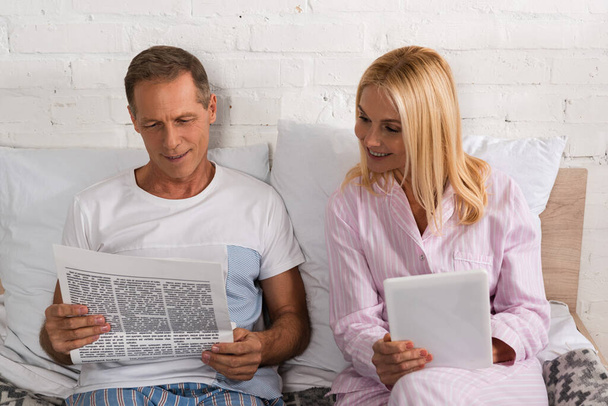 mature couple reading newspaper and using digital tablet on bed - Foto, afbeelding