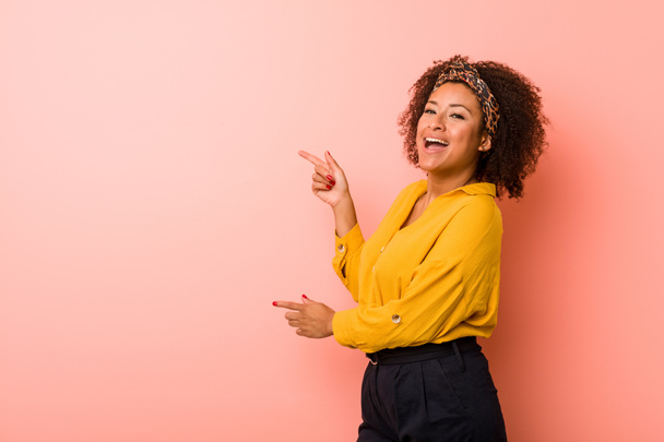 Joven mujer afroamericana sobre un fondo rosa excitado señalando con los dedos índice de distancia
. - Foto, imagen