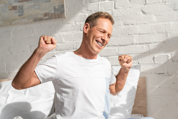 Smiling man stretching while sitting on bed at morning - Photo, Image