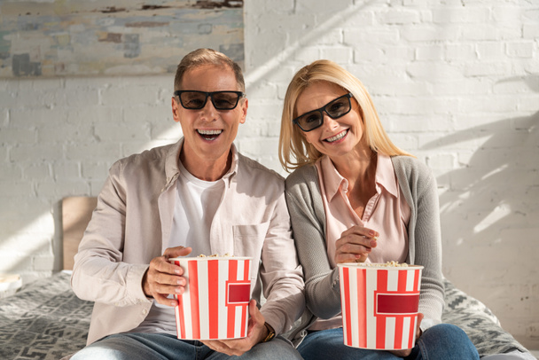 Smiling couple in 3d glasses holding buckets with popcorn on bed - Photo, image