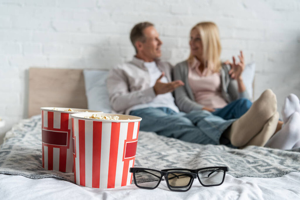 Selective focus of popcorn buckets and 3d glasses with talking couple on bed - Fotografie, Obrázek