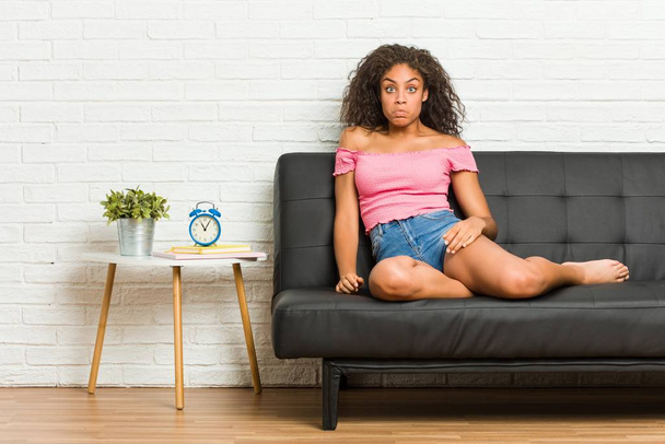 Młoda Afroamerykanka siedząca na kanapie wzrusza ramionami i oczy zdezorientowane. - Zdjęcie, obraz