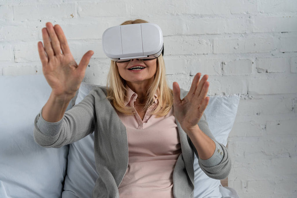 Lächelnde Frau im Virtual-Reality-Headset gestikuliert auf dem Bett - Foto, Bild