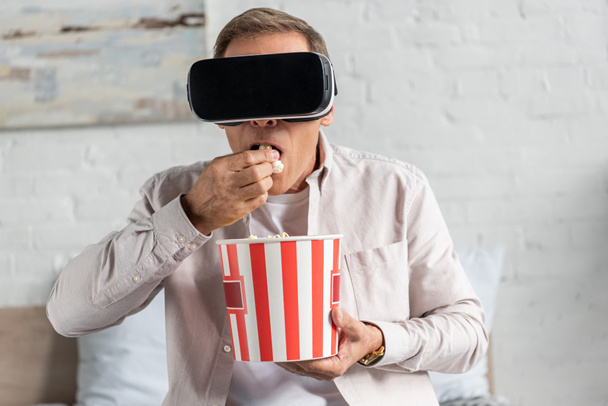 Man in virtual reality headset eating popcorn on bed - Фото, зображення