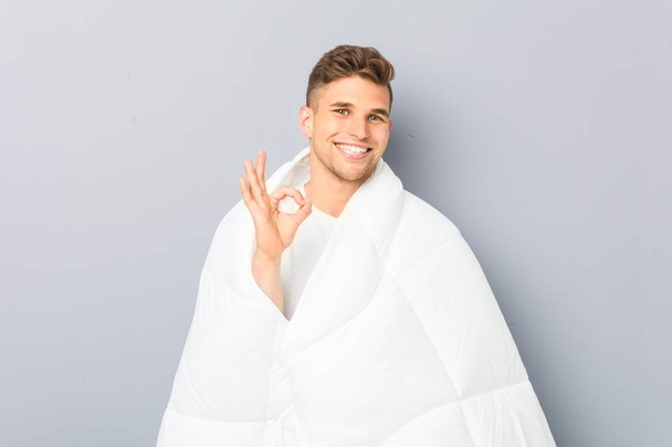Young man holding a nordic duvet cheerful and confident showing ok gesture. - Photo, Image