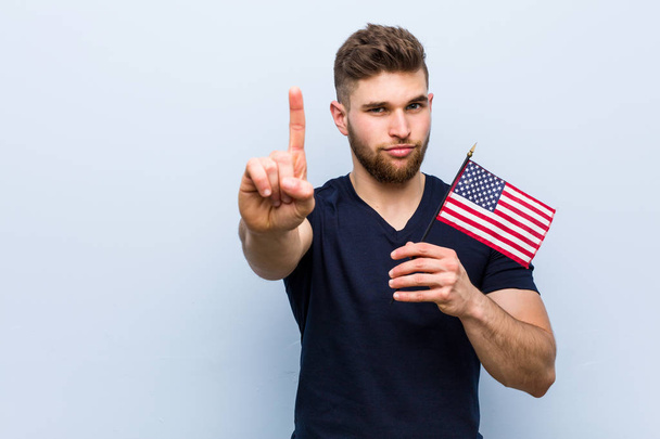 Amerika Birleşik Devletleri bayrağını tutan genç beyaz adam parmağıyla bir numarayı gösteriyor.. - Fotoğraf, Görsel