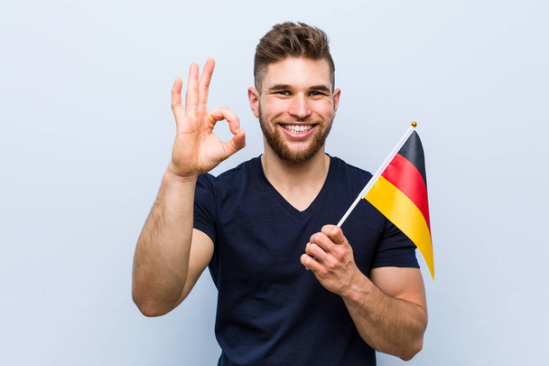 Jeune homme caucasien tenant un drapeau allemand joyeux et confiant montrant geste ok
. - Photo, image