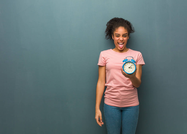 Young black woman funnny and friendly showing tongue. She is holding an alarm clock. - Foto, Bild