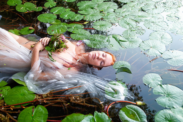 Giovane donna pazza infelice sexy signora Ophelia con i capelli rossi ricci si trova con i fiori con gli occhi chiusi morire in acqua nel lago
 - Foto, immagini