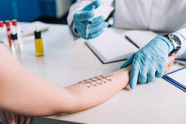 Ausgeschnittene Ansicht eines Allergologen in Latexhandschuhen, der eine Spritze in der Nähe einer Frau hält, während er einen Allergietest in der Klinik macht  - Foto, Bild