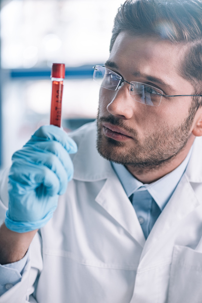 guapo inmunólogo barbudo en gafas mirando el tubo de ensayo con líquido rojo
  - Foto, Imagen