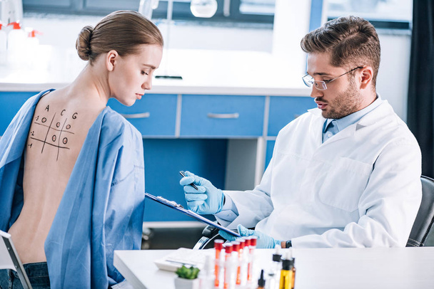selective focus of handsome allergist holding clipboard near woman with marked back  - Foto, imagen