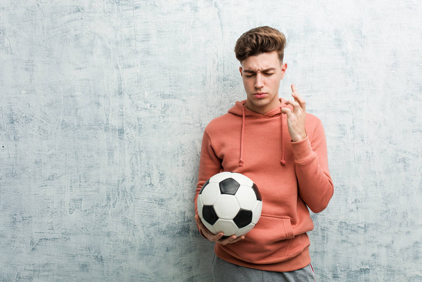 Νεαρός αθλητικός άνδρας κρατώντας μια μπάλα ποδοσφαίρου σταυρώνει τα δάχτυλα για την τύχη - Φωτογραφία, εικόνα