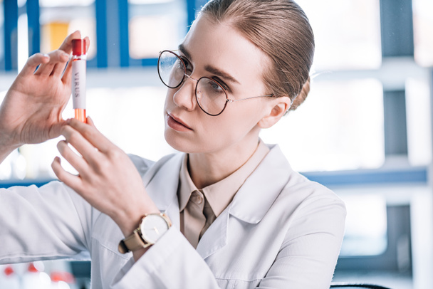 selective focus of beautiful immunologist in glasses looking at test tube with virus letters  - Photo, Image