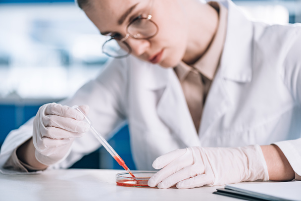 selective focus of attractive immunologist in eye glasses holding pipette with red liquid in laboratory  - Fotografie, Obrázek