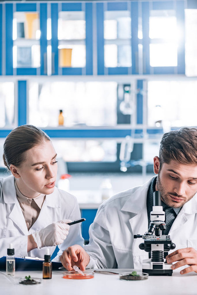 attractive biochemist looking at coworker near microscope  - Fotografie, Obrázek