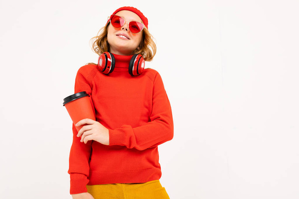 linda chica en gafas rojas posando con auriculares y sosteniendo taza de papel en el estudio sobre fondo claro
  - Foto, Imagen