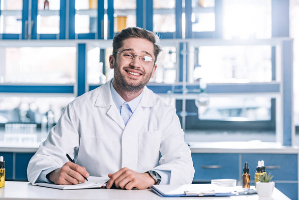 szczęśliwy człowiek w okularach i biały płaszcz siedzi przy biurku w klinice  - Zdjęcie, obraz