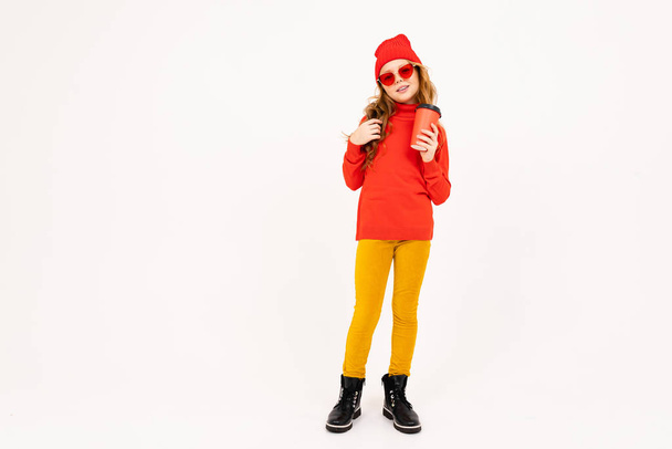 χαριτωμένο κορίτσι με κόκκινα γυαλιά που ποζάρουν με ακουστικά και κρατώντας χάρτινο κύπελλο στο στούντιο στο φως φόντο  - Φωτογραφία, εικόνα