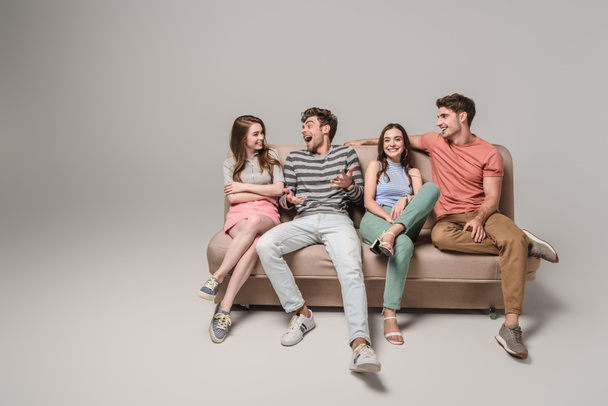 happy young friends talking and sitting on sofa on grey  - Φωτογραφία, εικόνα