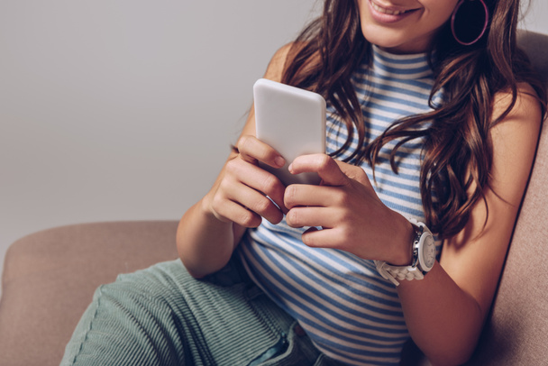 cropped view of smiling girl using smartphone while sitting on sofa isolated on grey - Fotó, kép