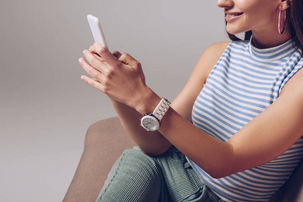 cropped view of smiling girl using smartphone while sitting on sofa isolated on grey - Foto, Imagem