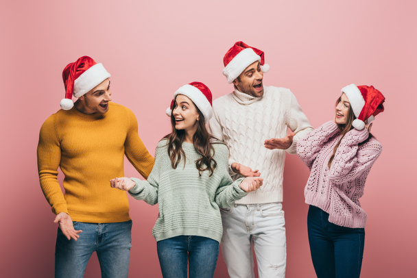surprised friends in sweaters and santa hats talking together, isolated on pink - Photo, Image